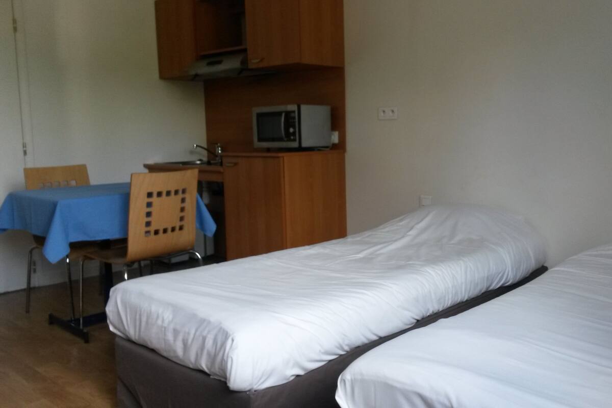 Ein Zimmer mit zwei Einzelbetten, einem Holztisch, einem Stuhl, einer Mikrowelle und einem Tisch mit blauer Tischdecke.