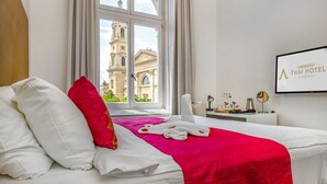 A hotel room with a bed, a window with a view of a historic building, a TV screen with the hotel name, and a small table with toiletries. - Andrassy Thai Hotel (Budapest)