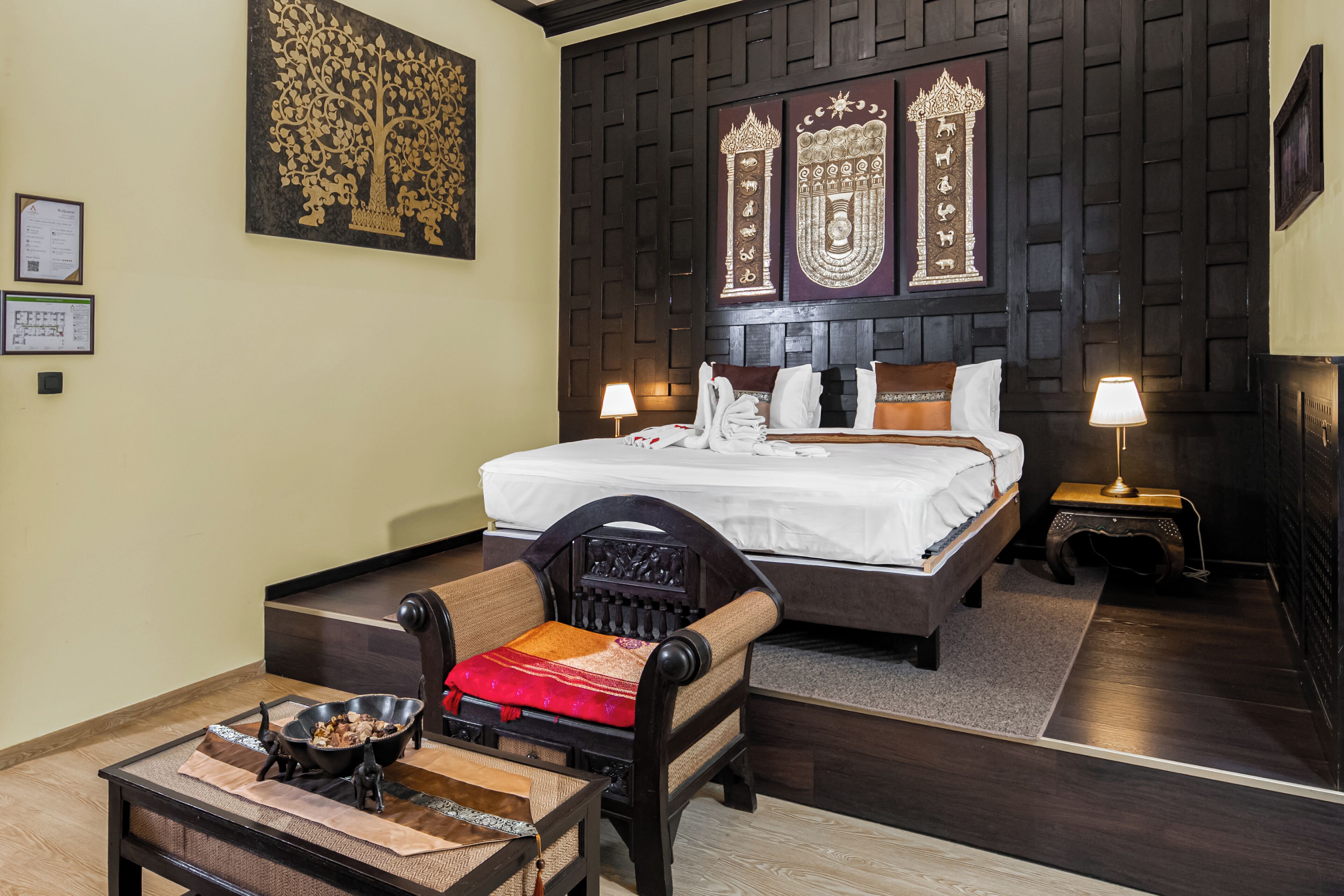 A hotel room with a bed, a chair, a table with a bowl and utensils, and wall decorations.