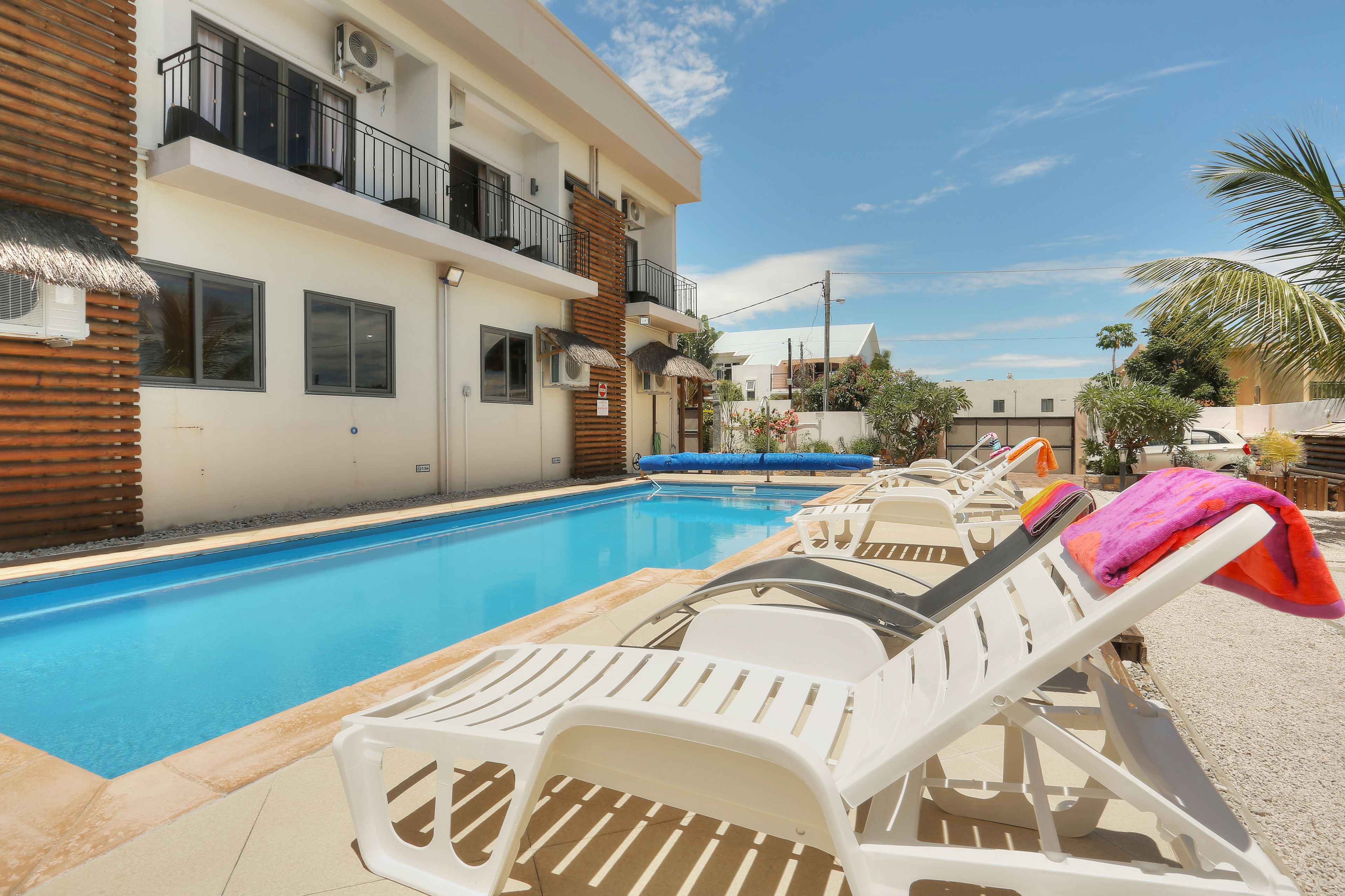 Una piscina amb gandules i ombrel·les, un edifici amb balcons, i un cel serè.