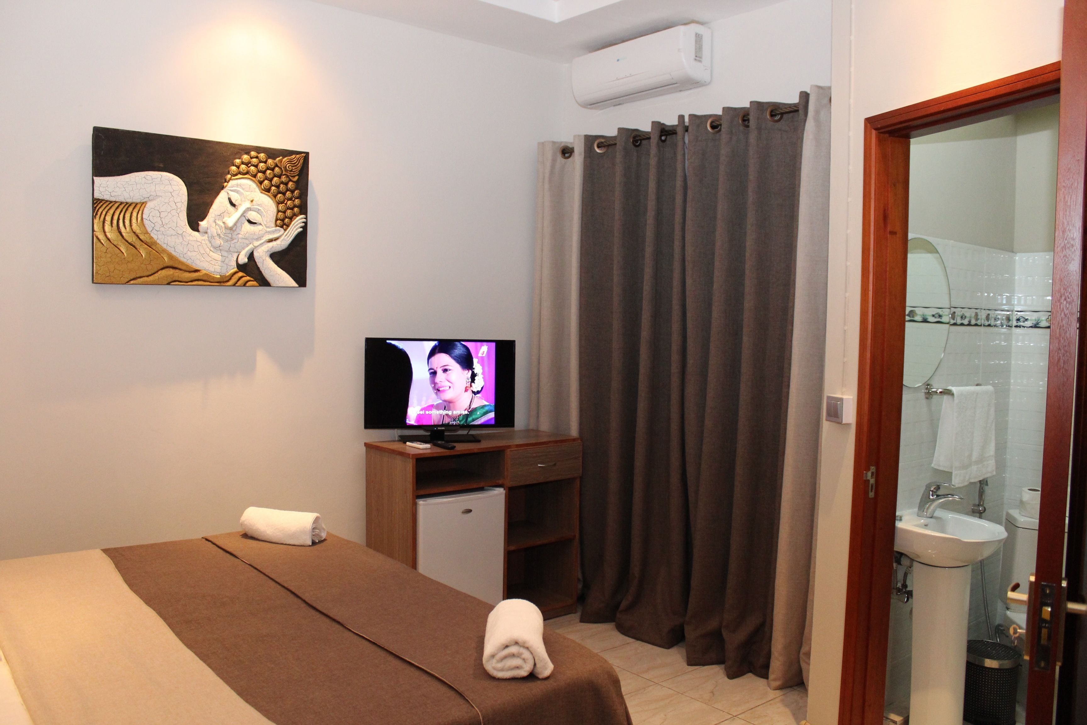 Una habitación de hotel con cama, televisión, un pequeño escritorio, baño con lavamanos e inodoro, y un cuadro en la pared.