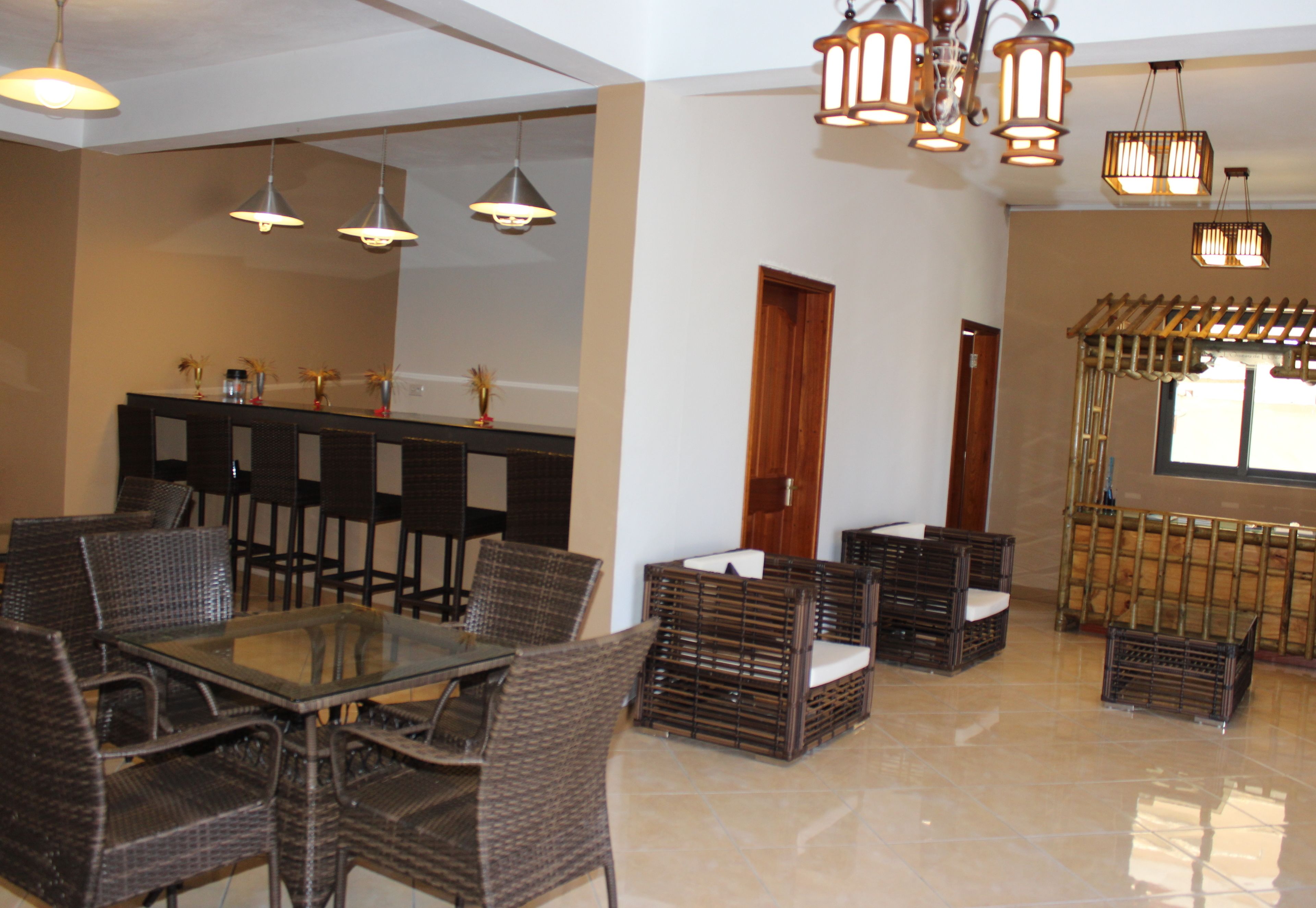 Amplo espaço interno com mesa de vidro cercada por cadeiras de vime, área de bar com banquetas e luminárias pendentes em estilo lanterna.
