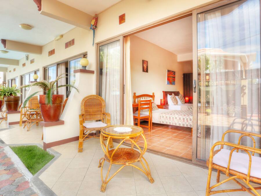 Ein Zimmer mit einem Bett, einem Sessel, einem Tisch und einer Schiebetür zu einem Balkon mit Blumentöpfen und einem Korbstuhl.