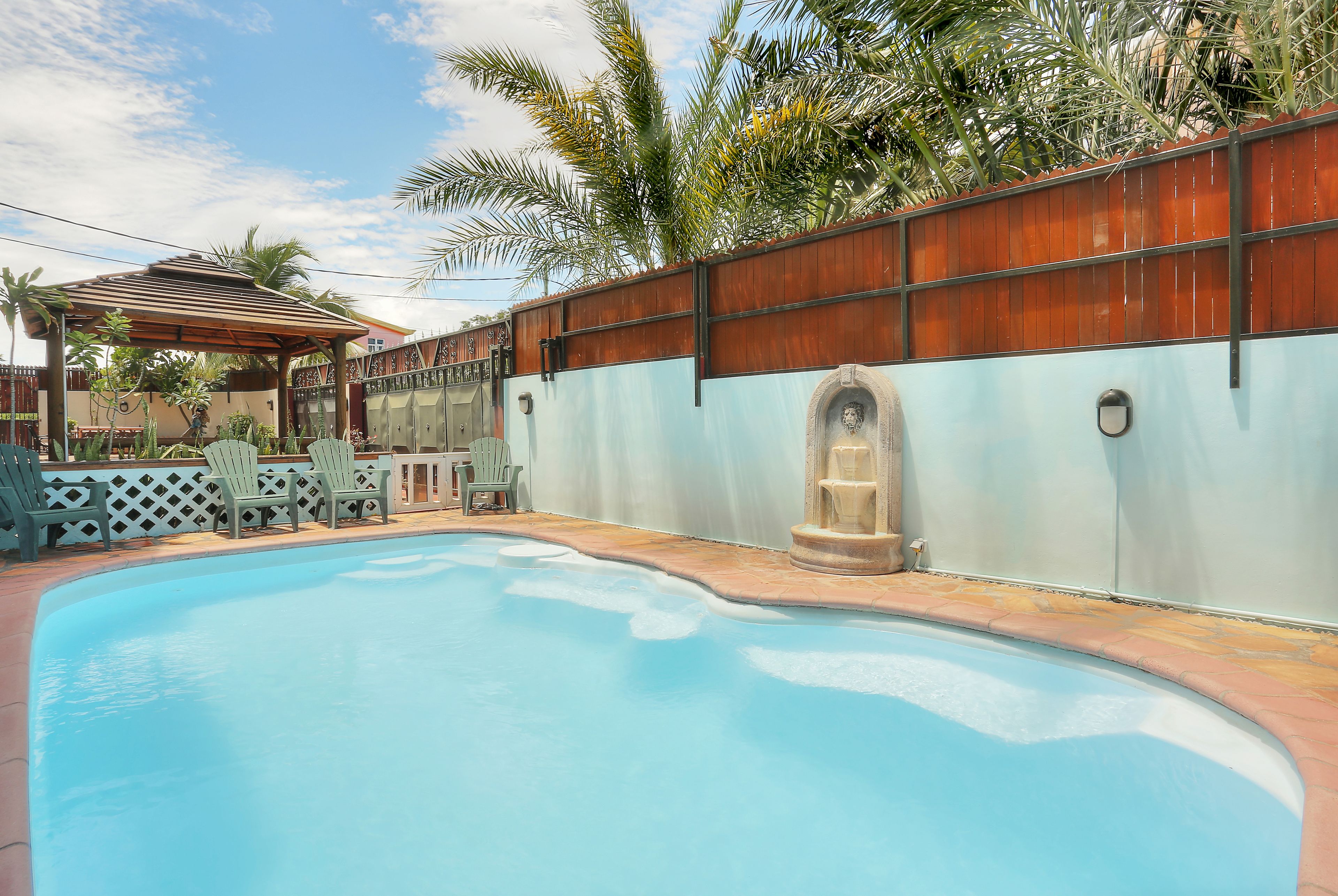 Una piscina con valla de madera, zona de estar cubierta y palmeras.