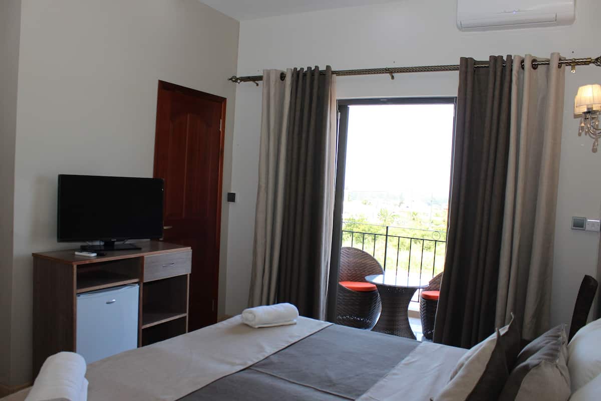Ein Hotelzimmer mit Bett, Fernseher, kleinem Kühlschrank, Balkon mit Tisch und Stuhl sowie Vorhängen.