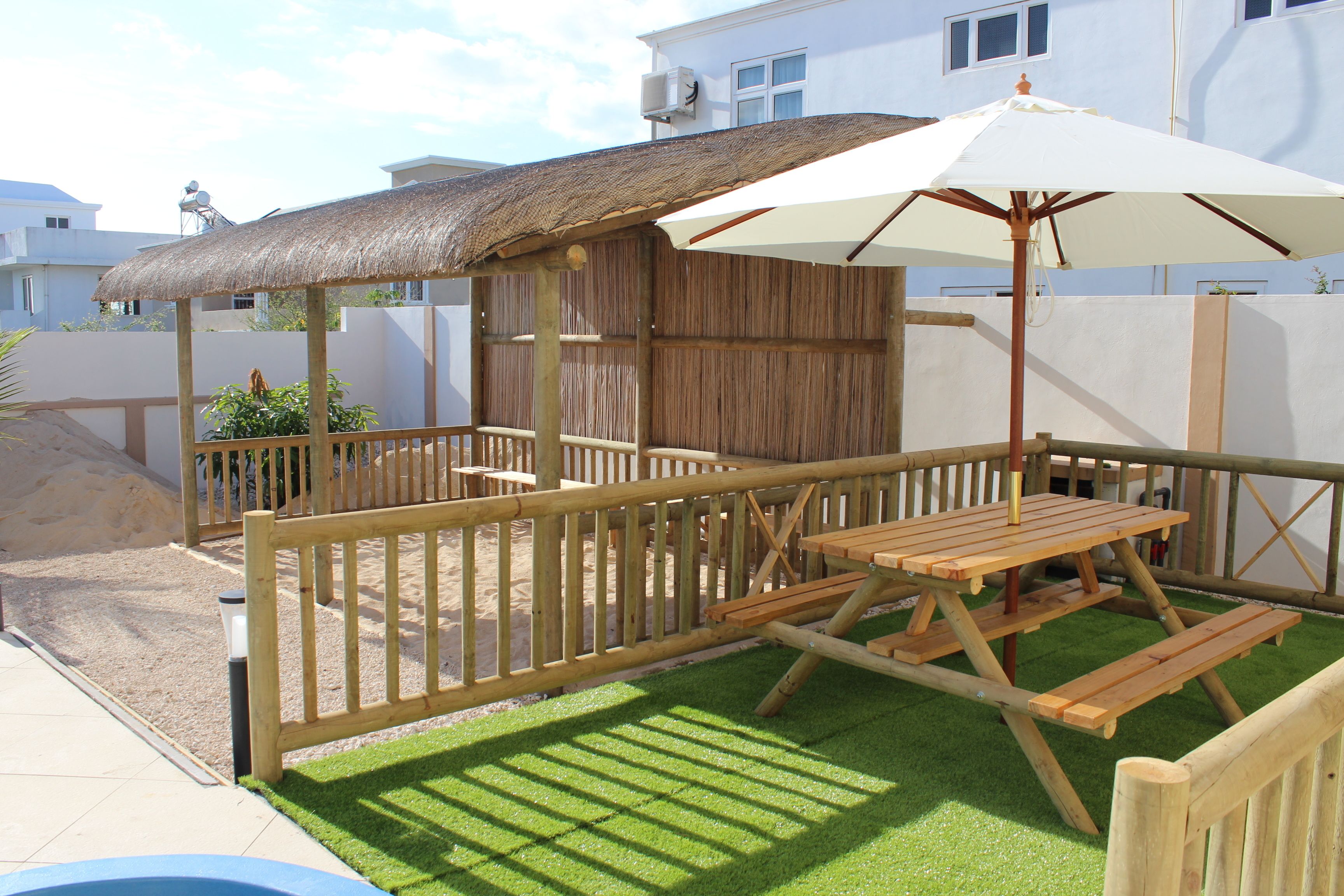 Une terrasse en bois surmontée d’un toit de chaume et agrémentée d’un parasol blanc, le tout entouré d’une clôture en bois.