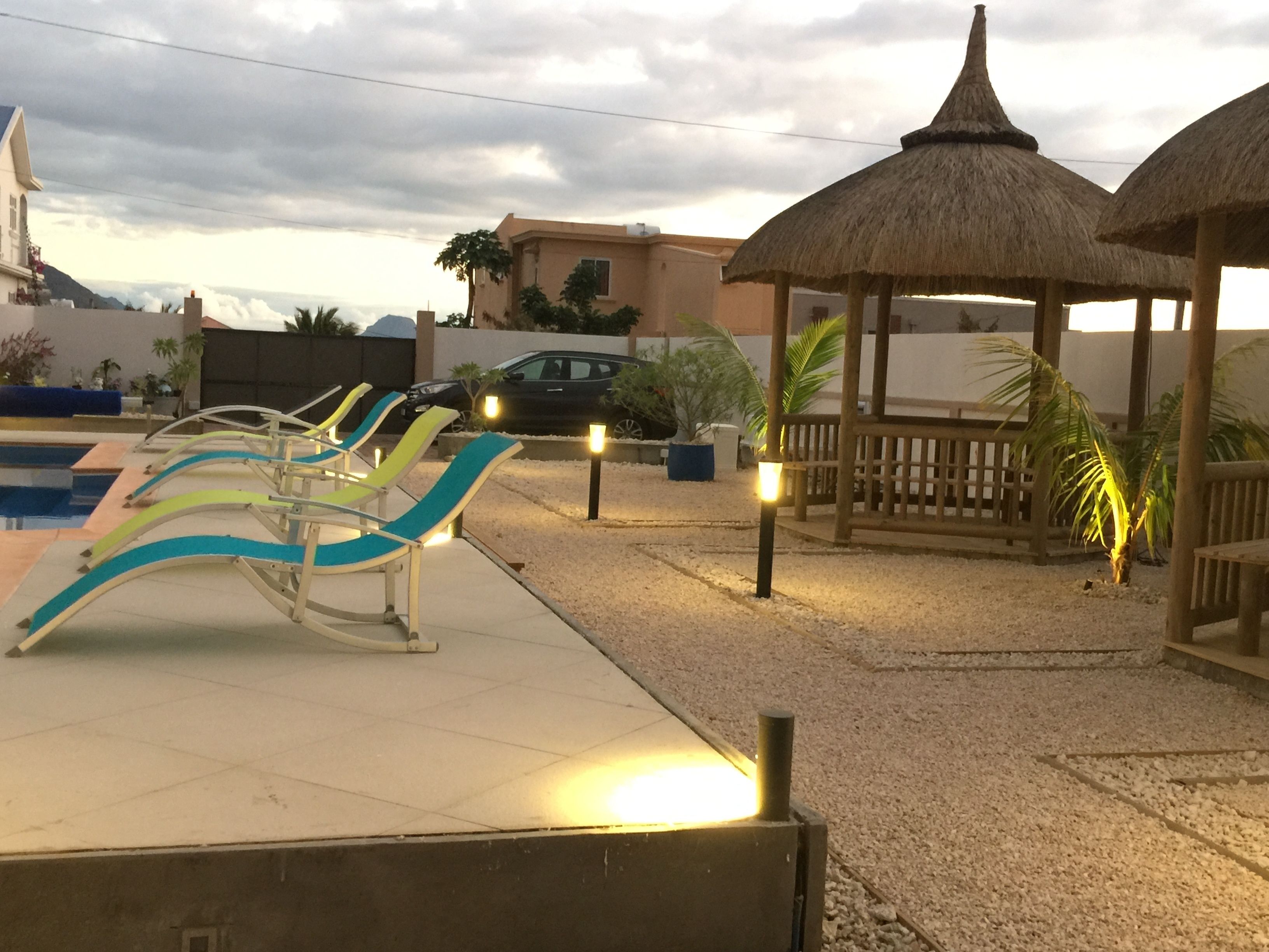 Un espace aménagé au bord de la piscine, avec des chaises longues, un gazebo à toit de chaume et une voiture garée à proximité.