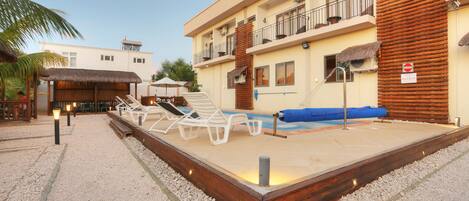 A pool area with lounge chairs, a thatched-roof structure, and a building with balconies.