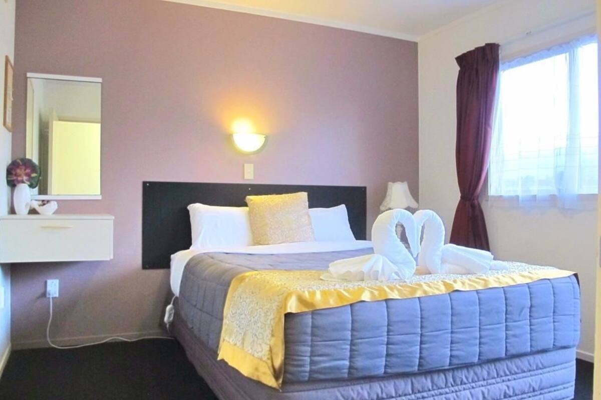 A hotel room with a bed, a nightstand, a wall-mounted lamp, a window with curtains, and a mirror.