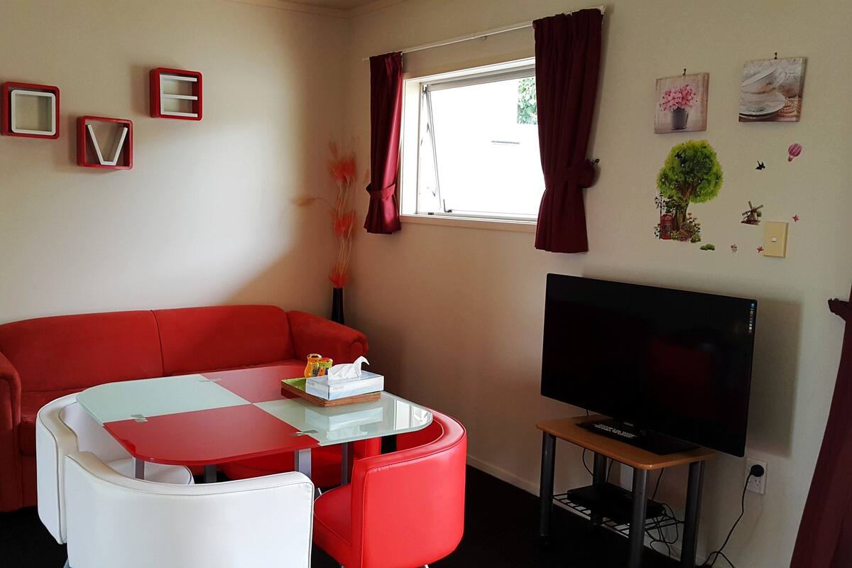 A compact living space with a red sofa, a small dining table, a TV on a stand, and a window with curtains.