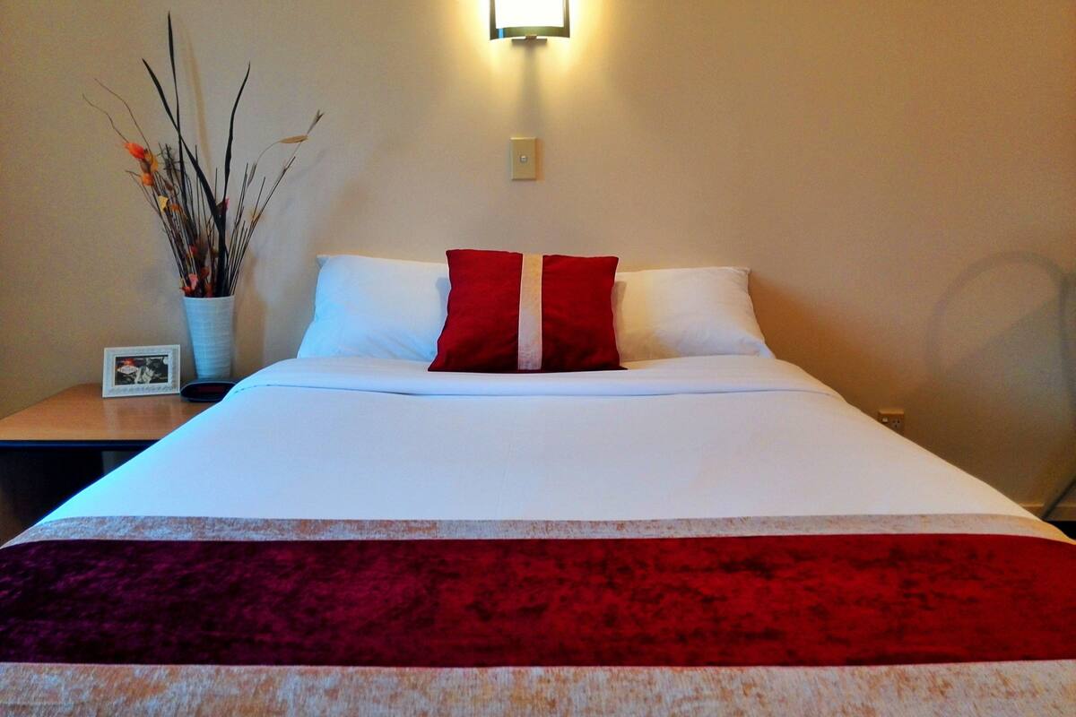 A neatly made bed with white and red pillows, a red throw blanket, and a vase with dried branches on a bedside table.
