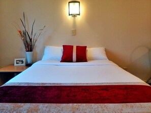 A neatly made bed with white and red pillows, a red throw blanket, and a vase with dried branches on a bedside table.
