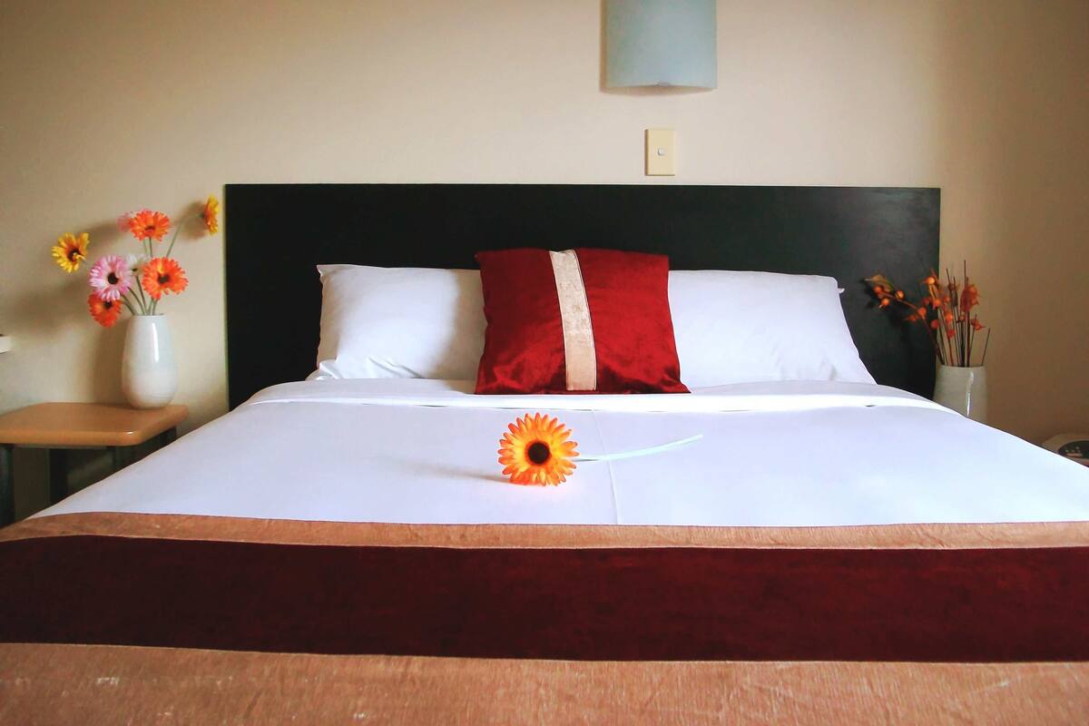 A neatly made bed with white and red bedding, a vase with flowers on the nightstand, and a wall-mounted light fixture.