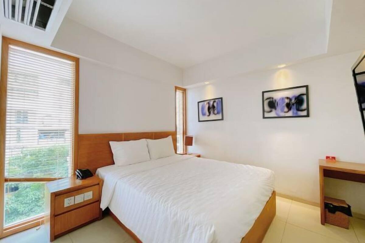 Een slaapkamer met een groot bed, een nachtkastje met een telefoon, een houten hoofdbord en een raam met jaloezieën.