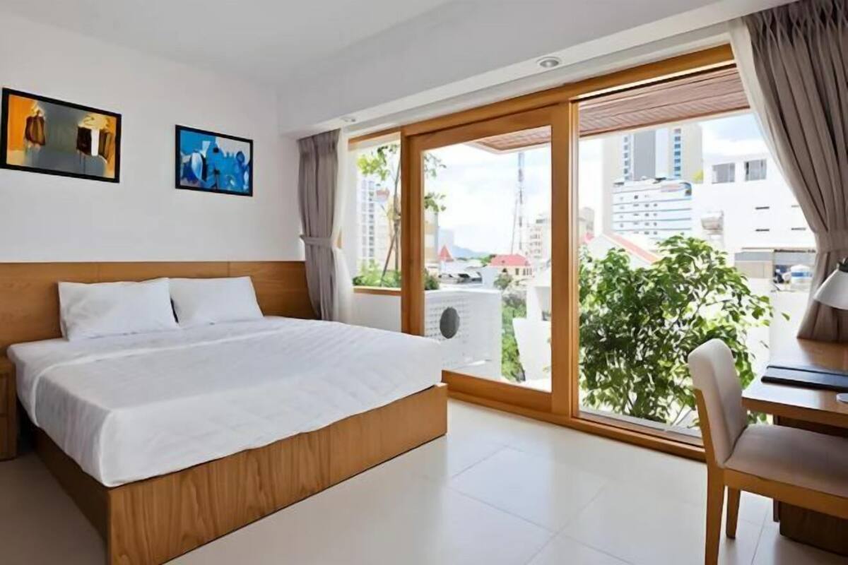 Een hotelkamer met een bed, een bureau en een groot raam met uitzicht op gebouwen en groen.
