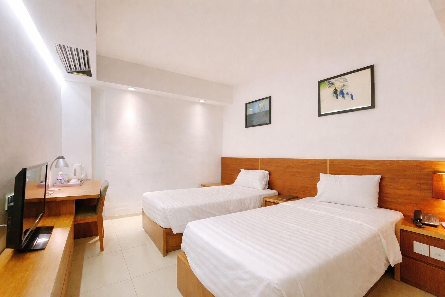 A hotel room with two single beds, a desk with a computer, a chair, a TV, and a small table with a lamp.