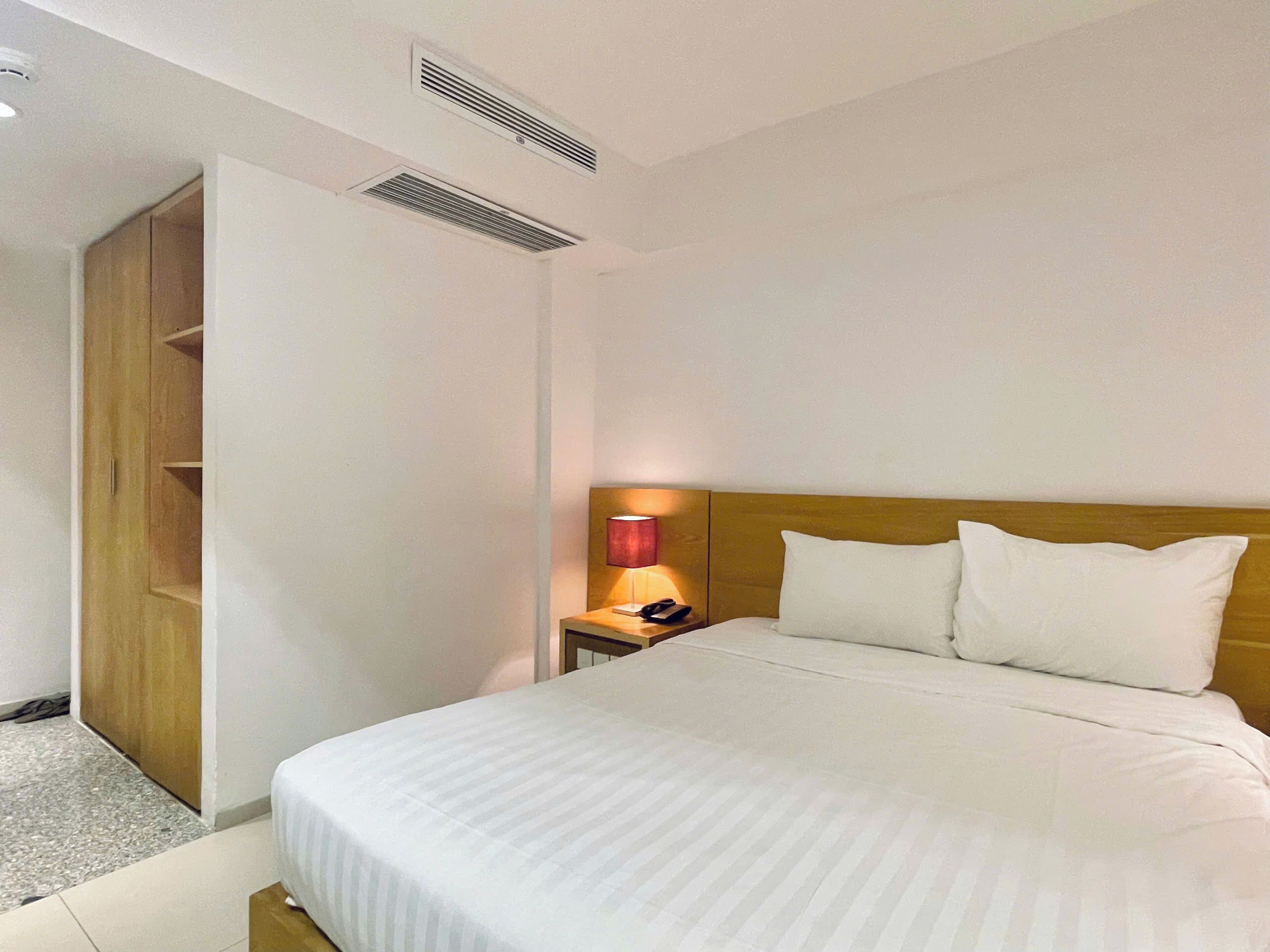 A hotel room with a bed, a bedside lamp, a wooden wardrobe, and a vent on the ceiling.