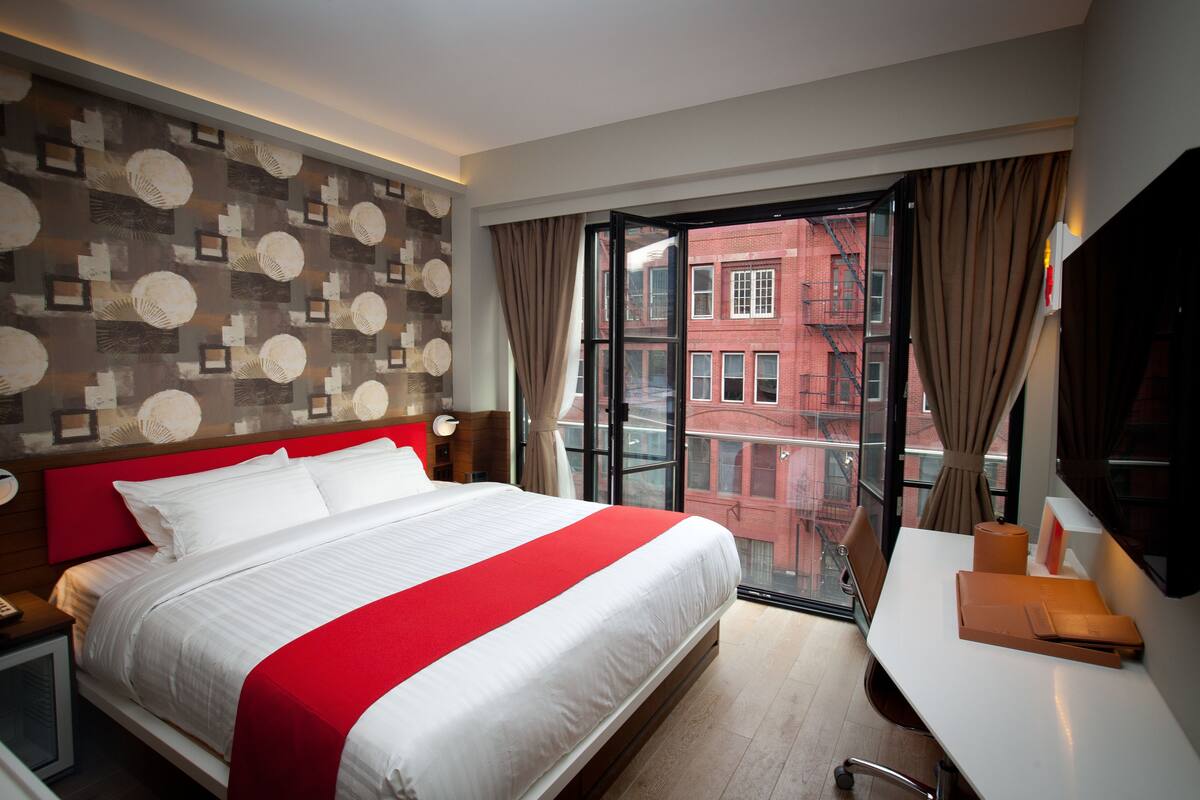 A hotel room with a large bed, a desk, and a view of a red brick building through the window.