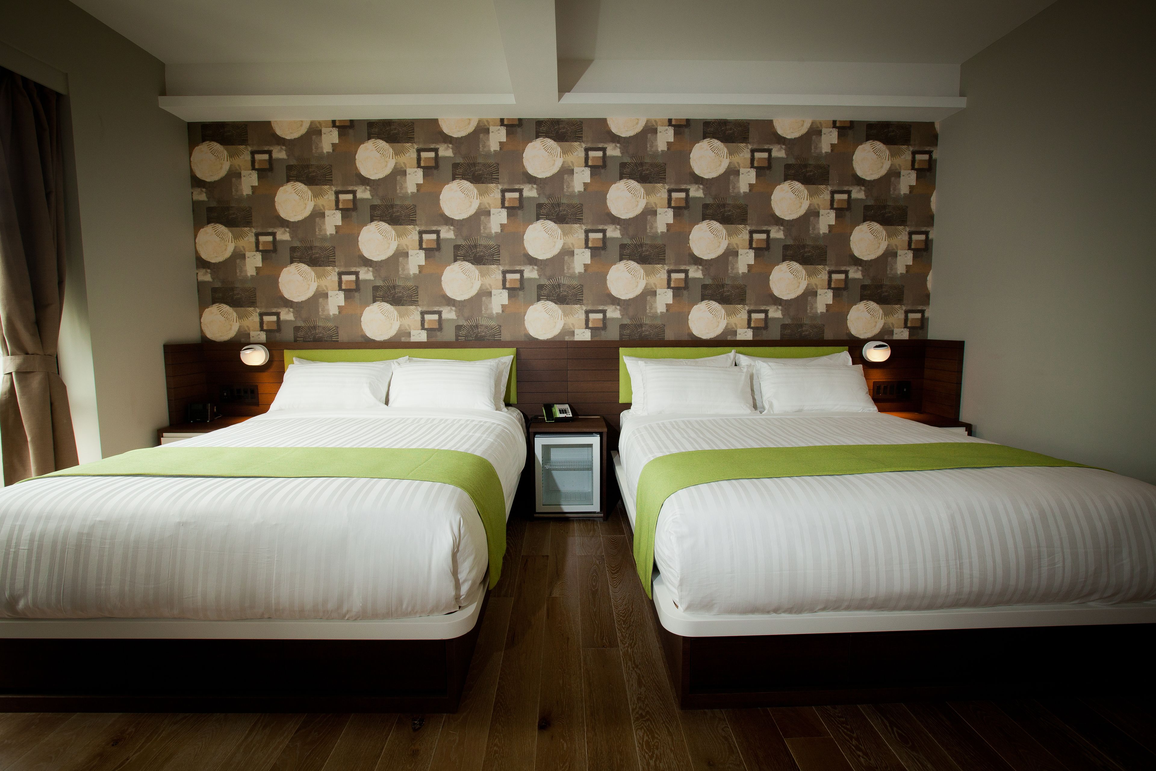 A hotel room with two beds, a patterned wall, and a small bedside table with a lamp.