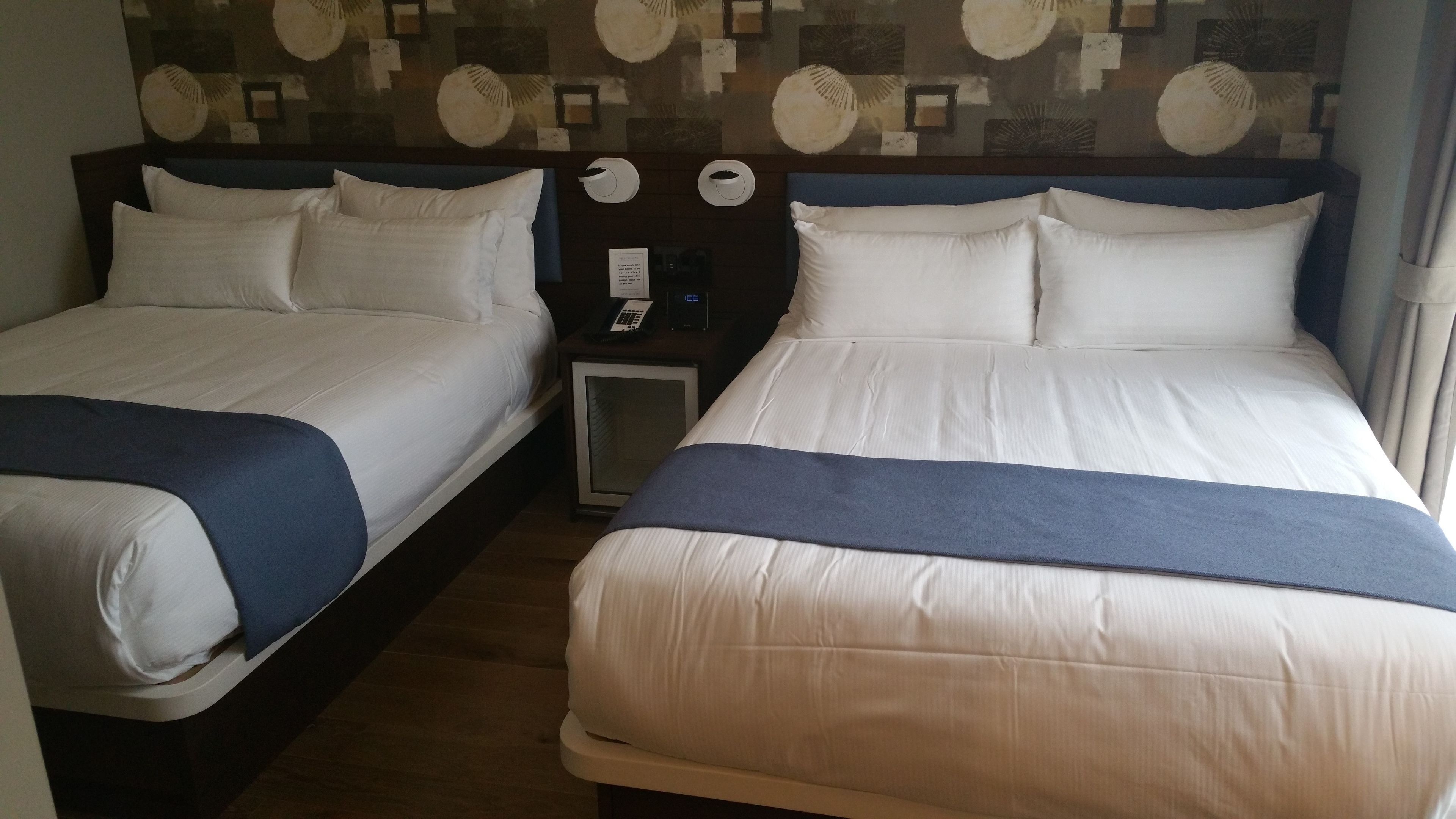 Een hotelkamer met twee bedden, elk voorzien van witte lakens en een blauwe accentkleur. Er staat een nachtkastje met een telefoon en een digitale klok.