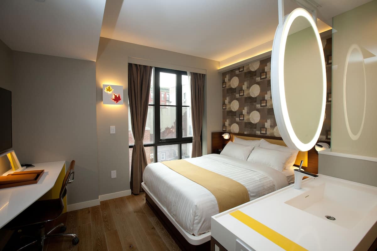 A hotel room with a bed, bedside table, desk, chair, and a large mirror.