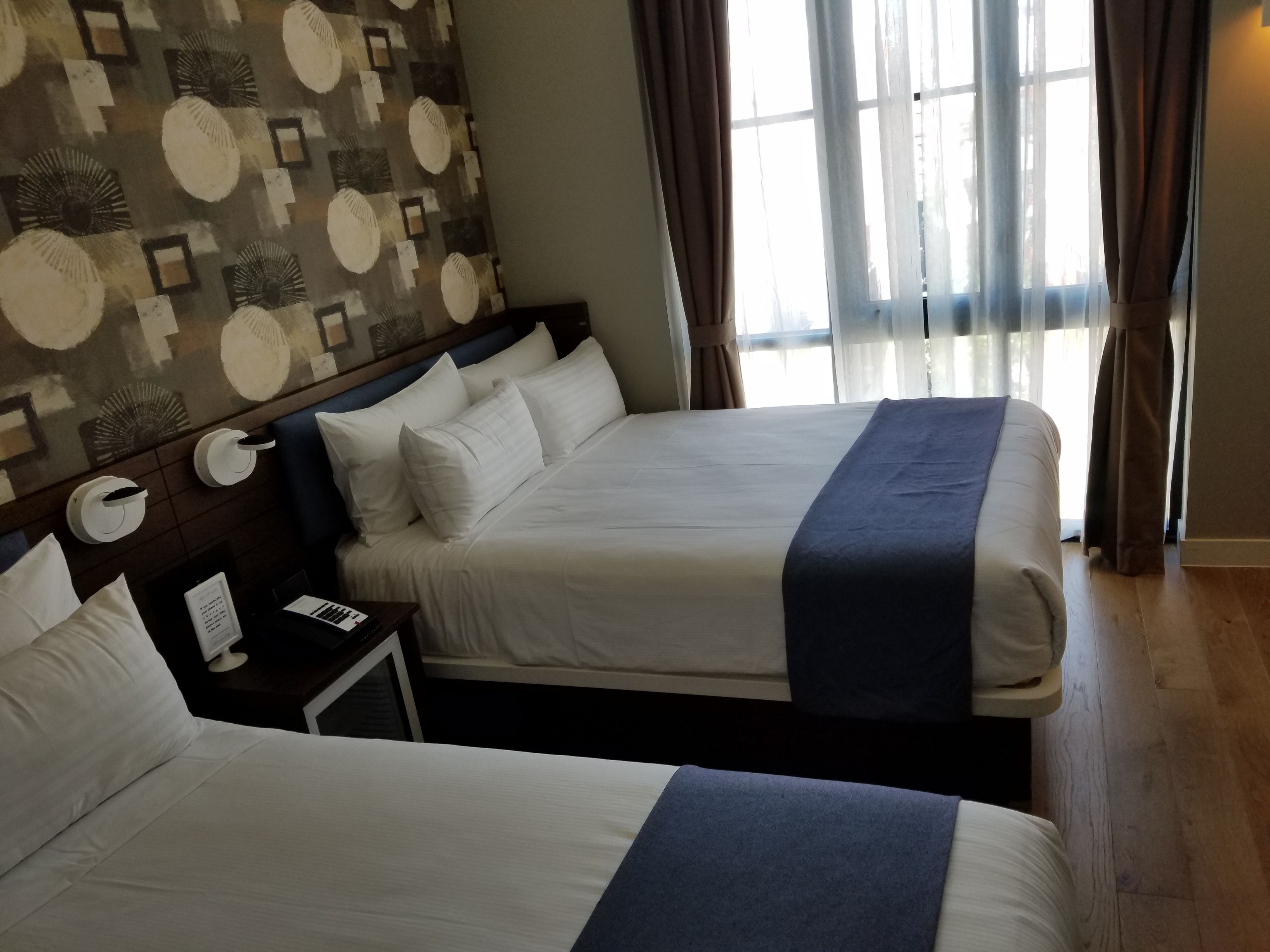 A hotel room with two beds, a nightstand, a lamp, and a window with curtains.