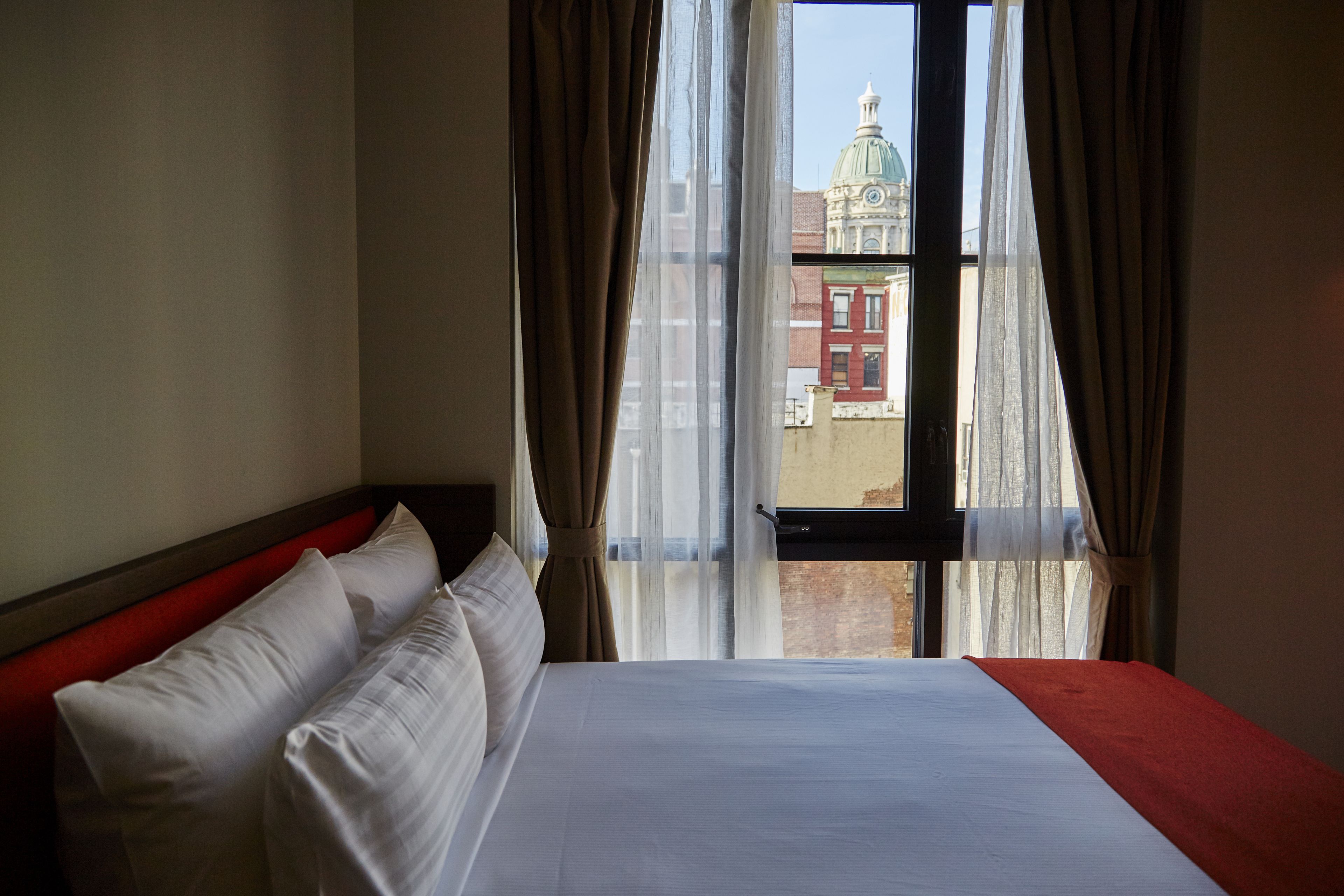 Habitación de hotel con una cama grande, cabecera roja y vistas a un edificio con cúpula a través de cortinas translúcidas.