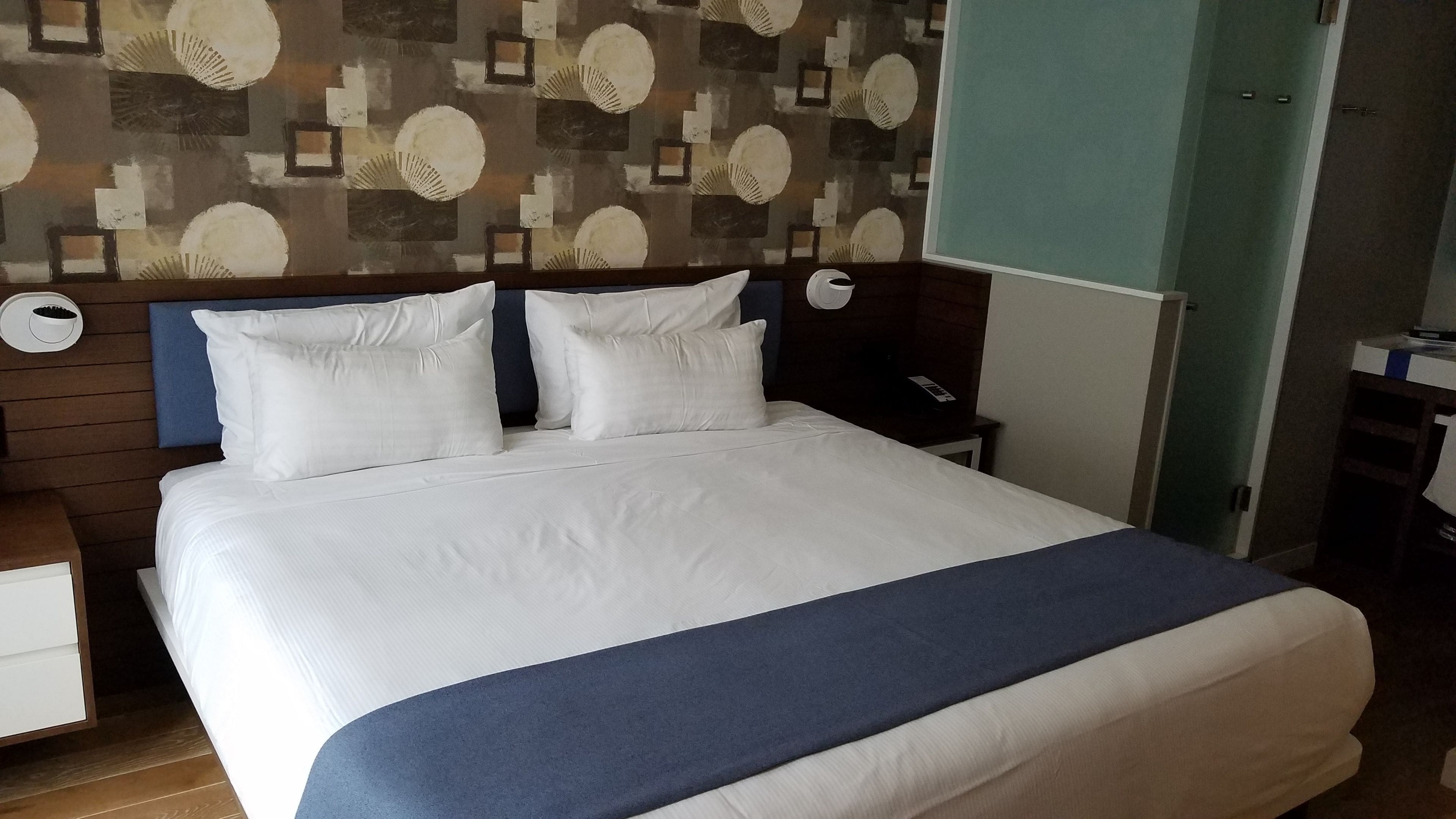 Una cama bien hecha con ropa de cama blanca y azul, un cabecero de madera y un papel tapiz estampado detrás.