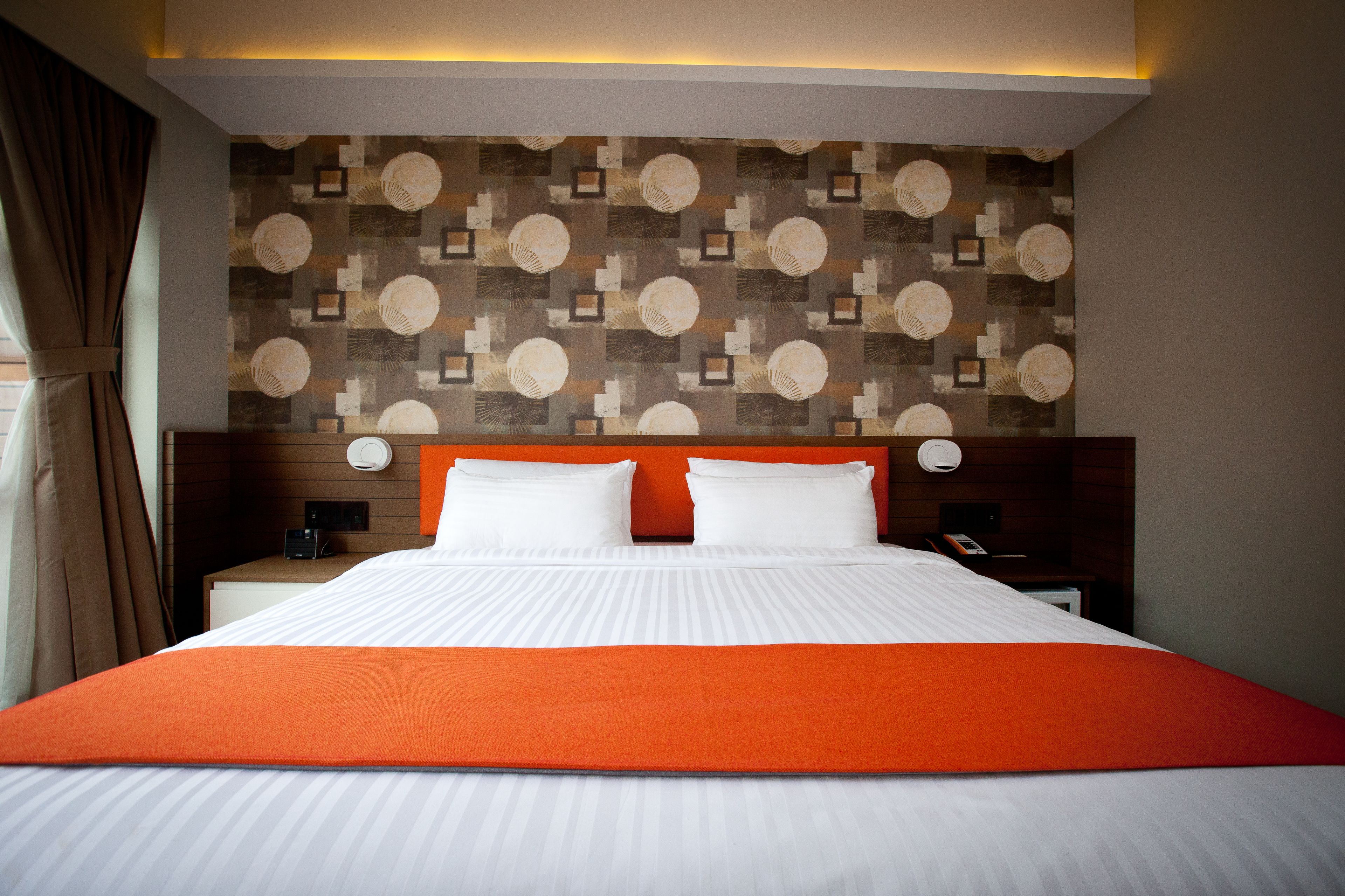 A hotel room with a large bed, two bedside tables, a headboard with a geometric pattern, and a window with curtains.
