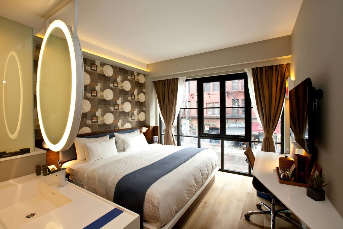 A hotel room with a large bed, a desk, a chair, a desk lamp, a mirror, and a window with curtains.