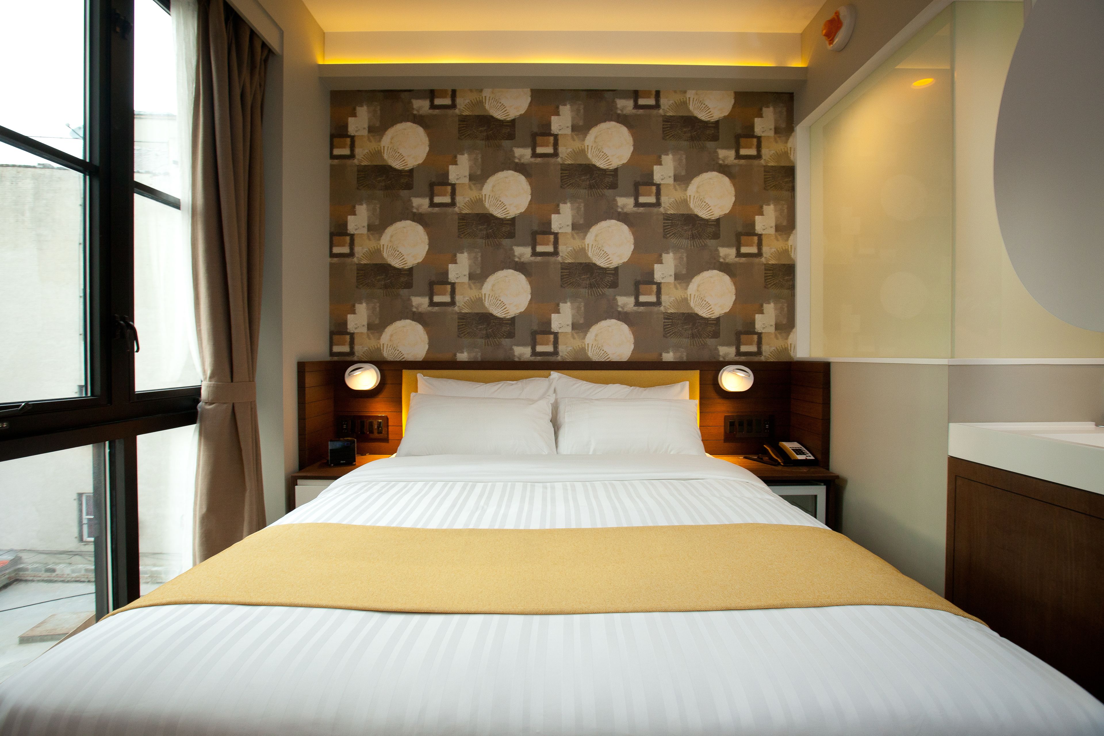 A hotel room with a large bed, a headboard with a patterned design, bedside lamps, and a view through a window.
