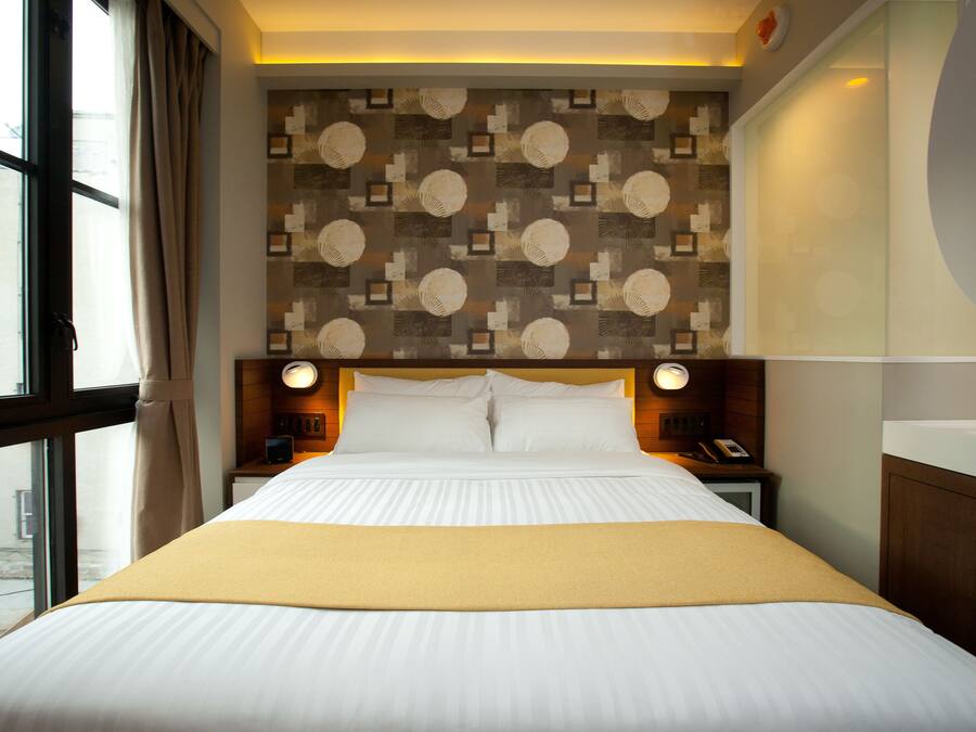 A hotel room with a large bed, a headboard with a patterned design, bedside lamps, and a view through a window.