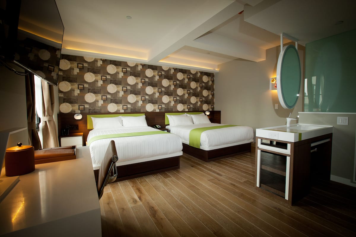 A hotel room with two beds, a bedside table, a small desk, a mirror, and a bathroom with a sink.