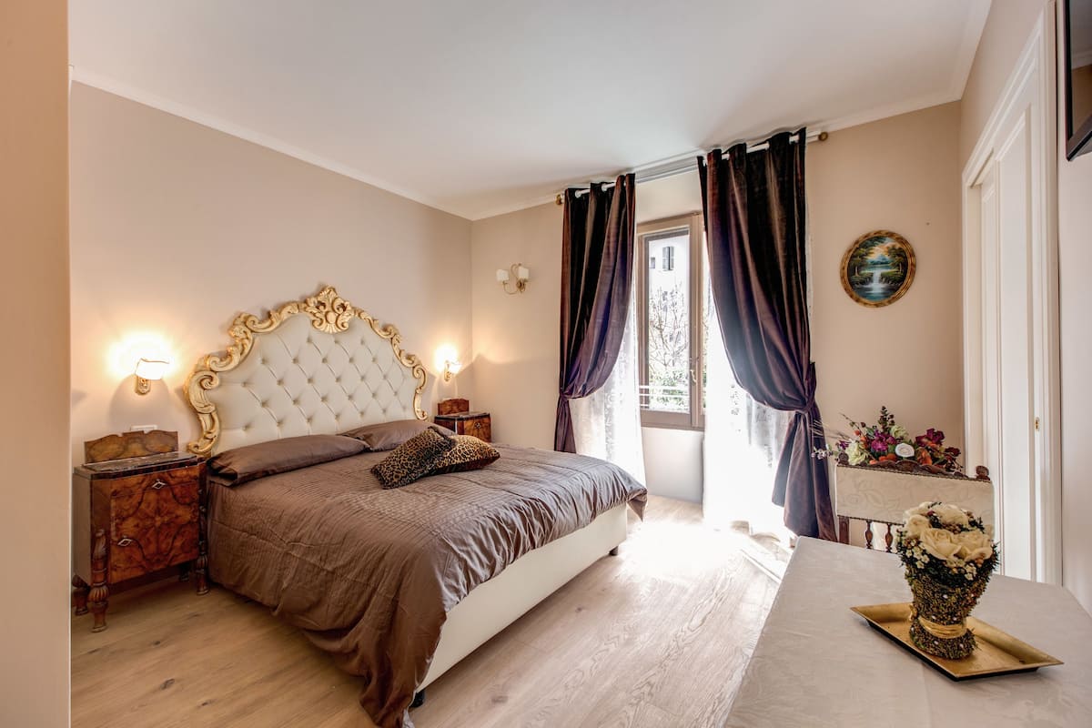 A bedroom with a tufted headboard, a wooden nightstand, a bedside table with a vase of flowers, and a view of the outdoors through a window with curtains.