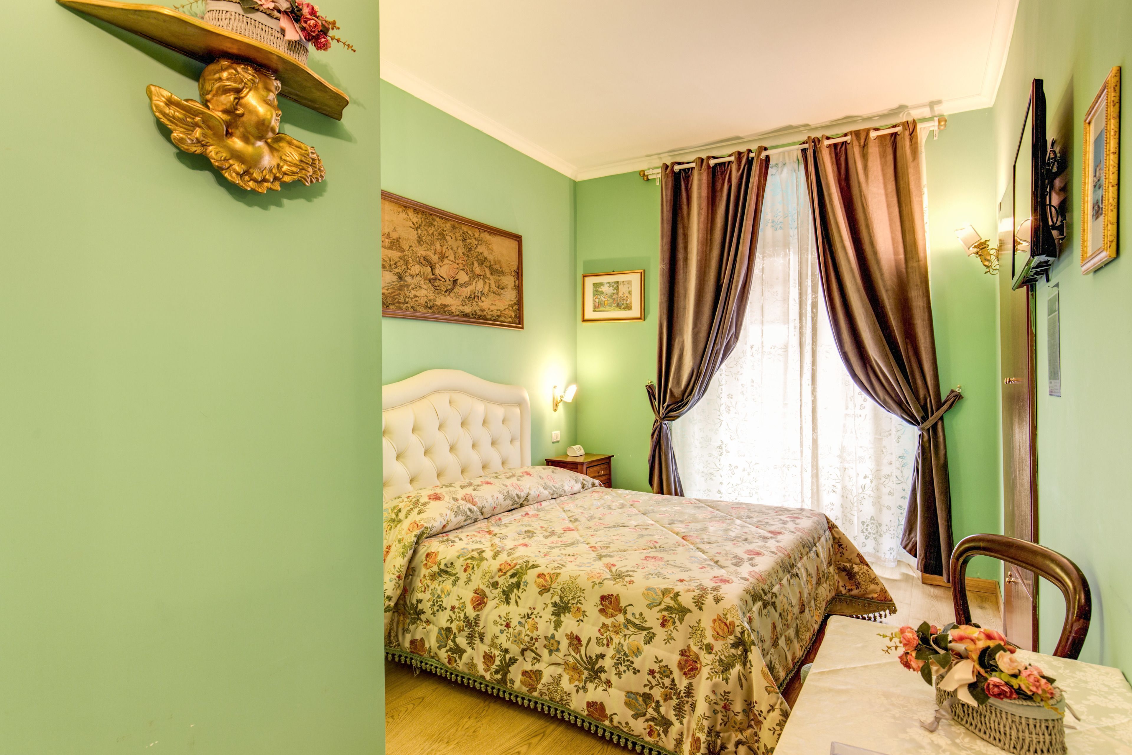 Ein Schlafzimmer mit Blumenmuster-Bettdecke, einem gepolsterten Kopfteil, einem Nachttisch, einer Lampe, einem Fenster mit Vorhängen und einem Wandregal mit einer Dekoration.