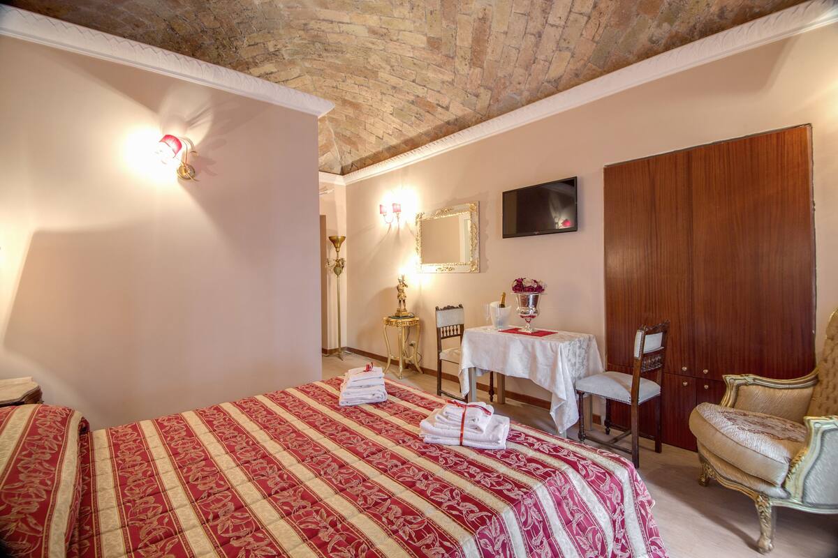 A hotel room with a bed, a wooden wardrobe, a chair, and a small table with a vase of flowers.