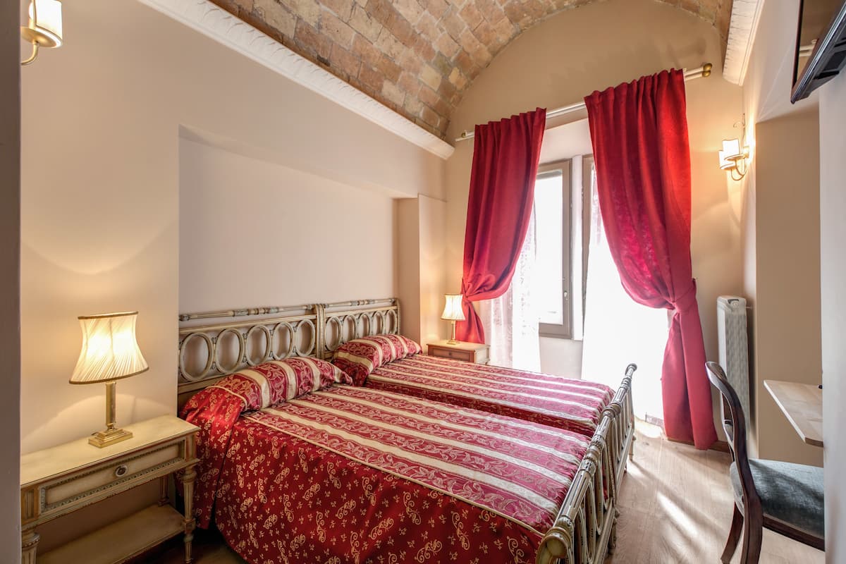 A room with two beds, a nightstand with a lamp, a wooden chair, and a window with red curtains.