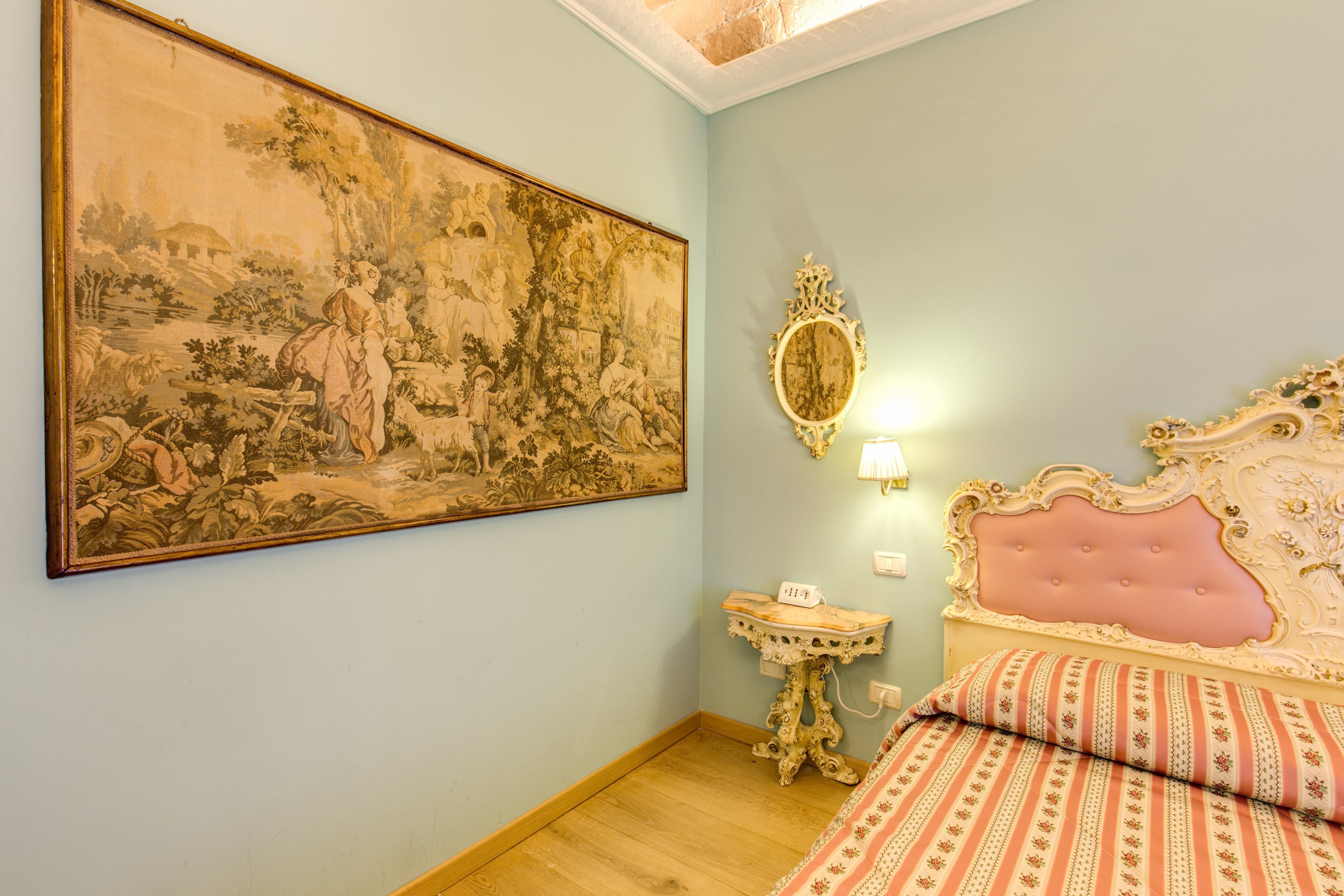A bedroom with a large landscape painting, a bedside table with a lamp, and a bed with ornate headboard and patterned bedding.