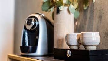 En kaffemaskine på en træbordplade med to keramikkrus og en vase med grønne blade.