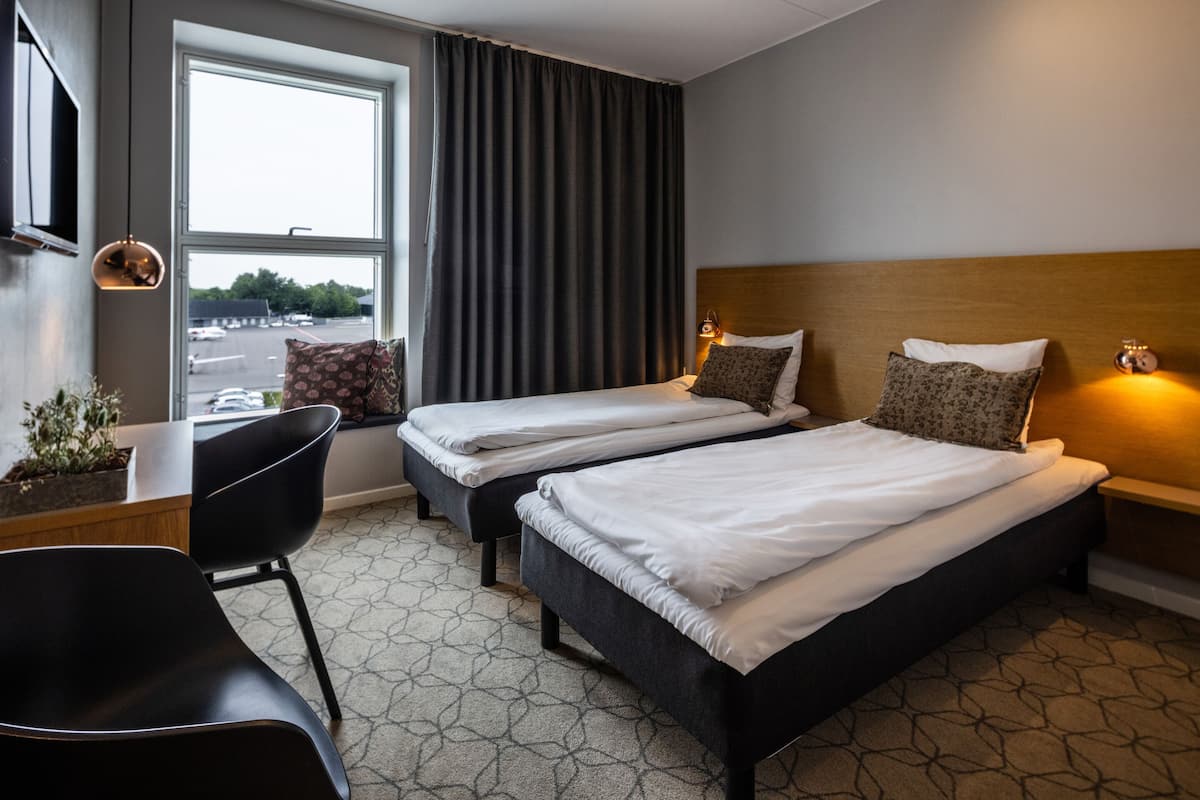 Ein Hotelzimmer mit zwei Betten, einem Schreibtisch, einem Stuhl und einem großen Fenster mit Vorhängen.
