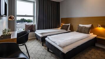 Quarto de hotel com duas camas, uma escrivaninha, uma cadeira e uma janela grande com cortinas.
