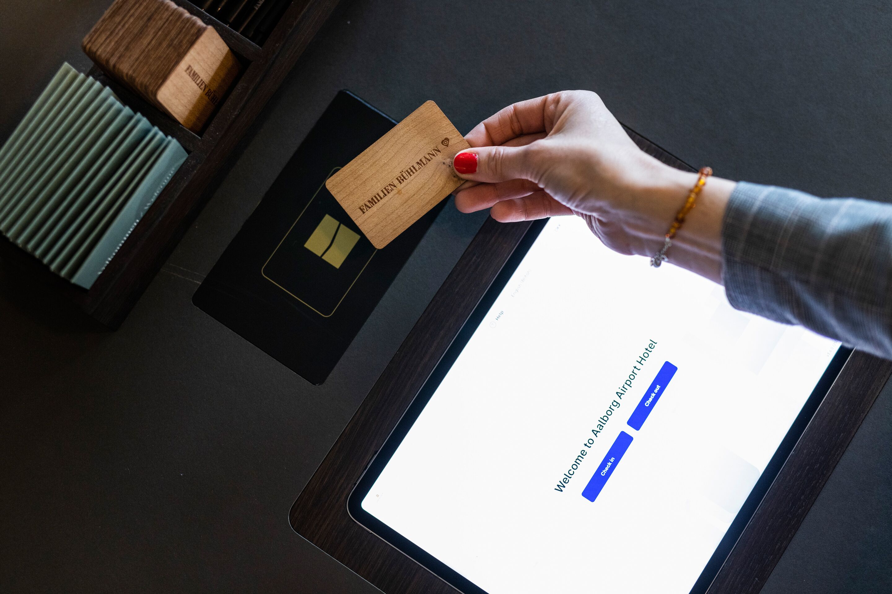 A hand holding a wooden card with "FAMILIEN RÜHL MANN" engraved on it, placed on a tablet displaying a hotel welcome page.