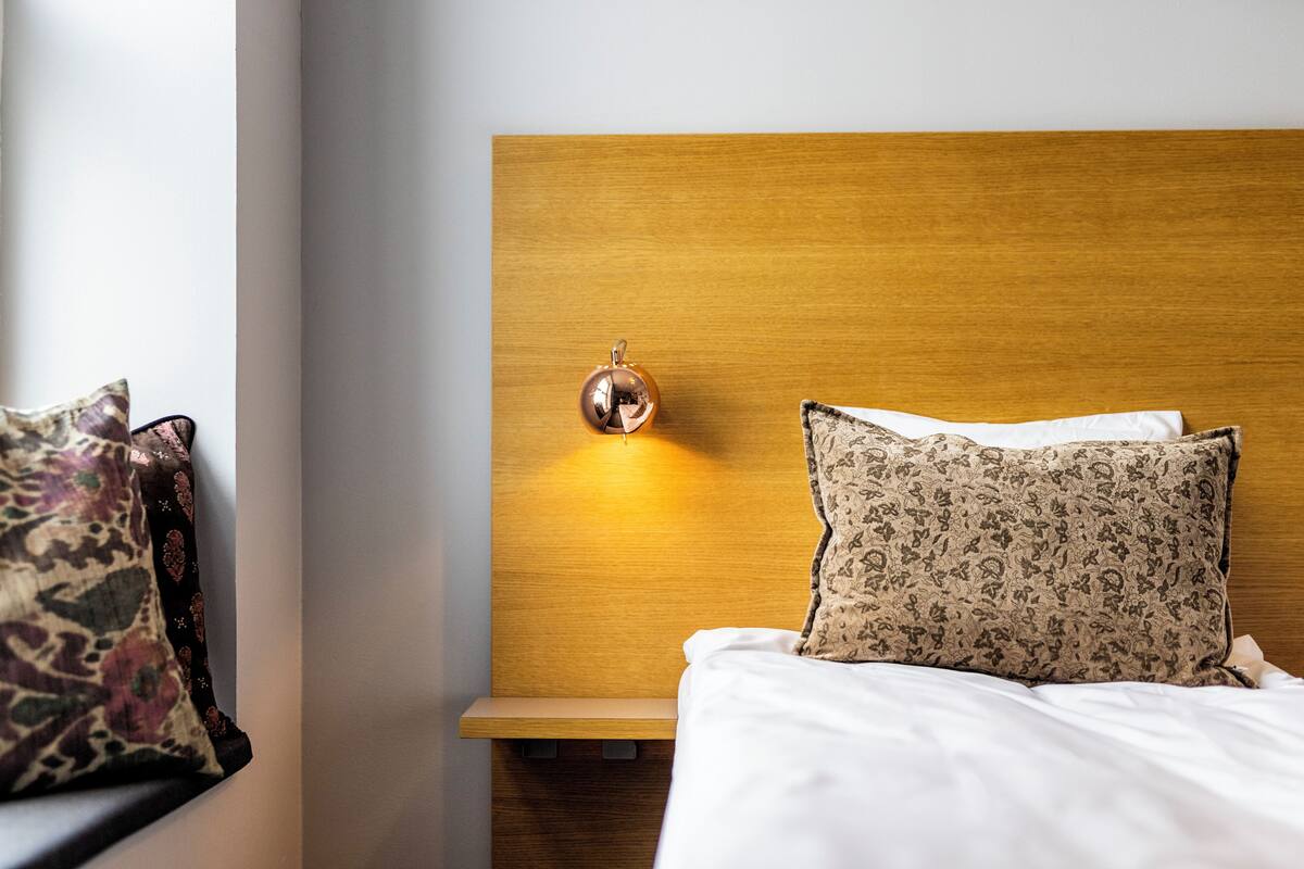 Ein ordentlich bezogenes Bett mit einem gemusterten und einem schlichten Kopfkissen, ein Nachttisch mit einer Dekoration sowie eine wandmontierte Lampe.