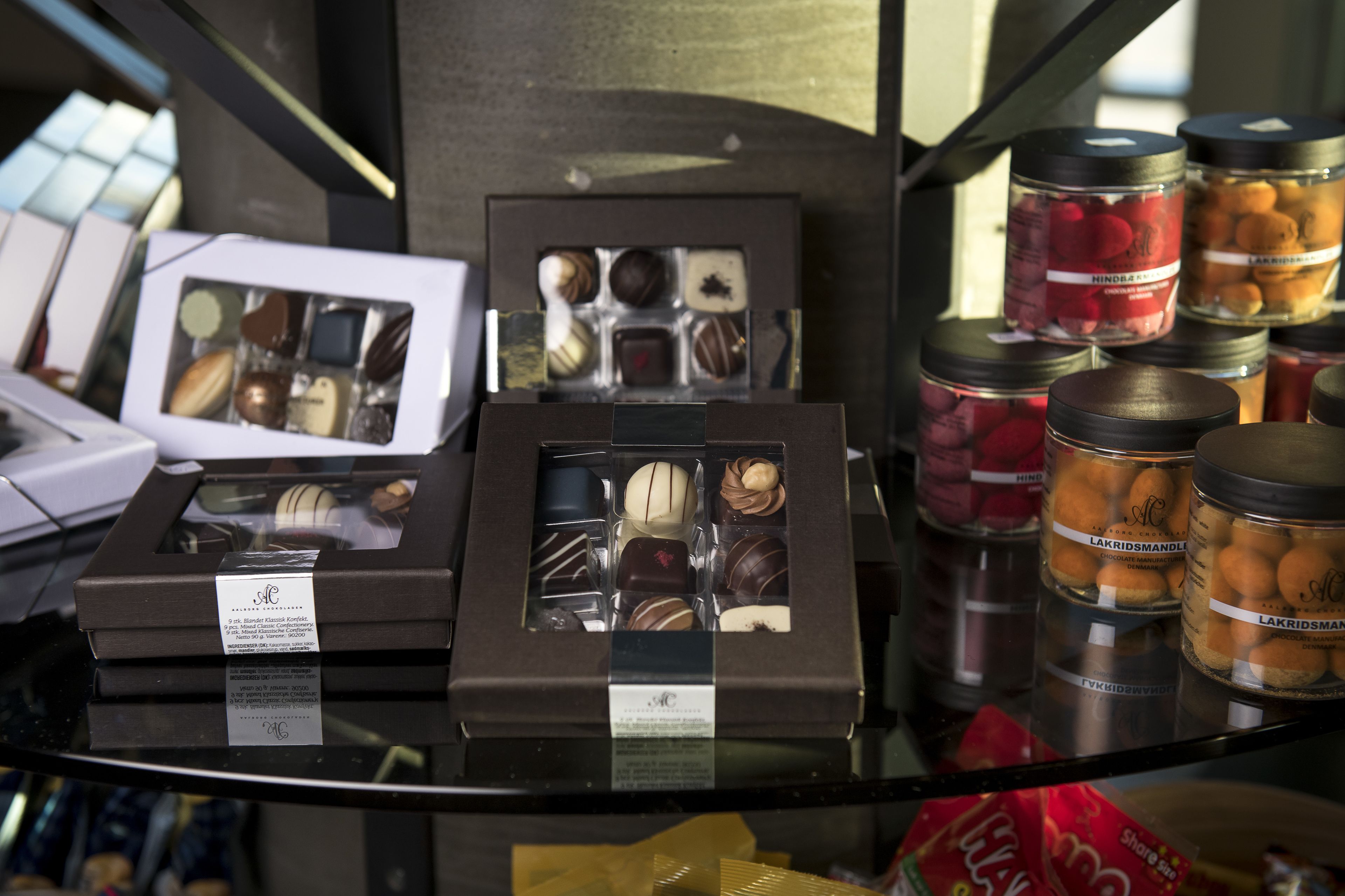 Una exhibición de bombones y dulces variados en diferentes envases, incluyendo cajas y frascos, sobre un estante de vidrio.