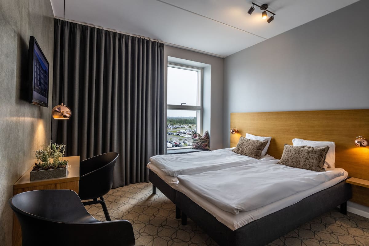Ein modernes Hotelzimmer mit einem großen Bett, einem Schreibtisch, einem Stuhl und Blick auf die Stadt durch ein Fenster.