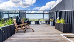 Une terrasse sur le toit, avec des chaises en bois et des jardinières noires, donnant sur un parking et un ciel dégagé.