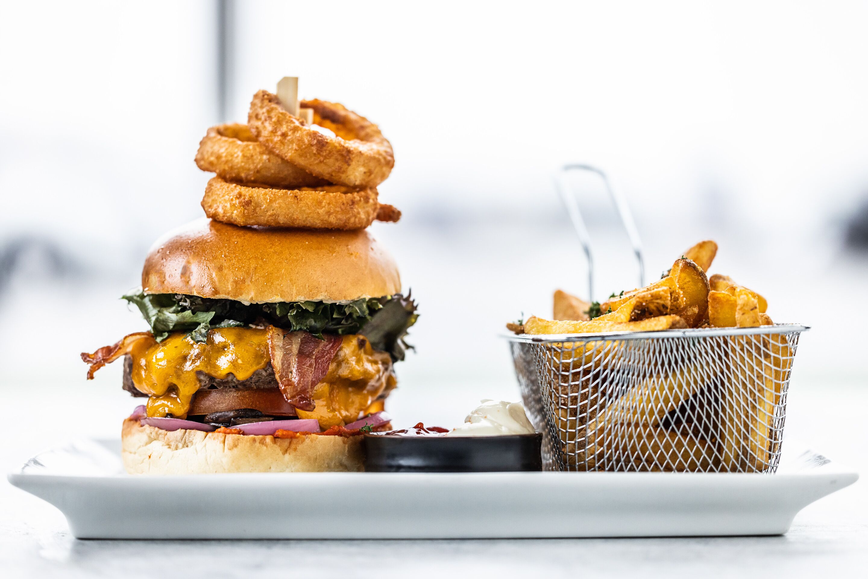 A burger with bacon, cheese, and lettuce, accompanied by onion rings and a side of fries.