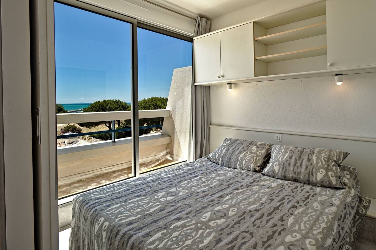 A bedroom with a large window, a bed with patterned bedding, a white wardrobe, and a view of the beach and sea.