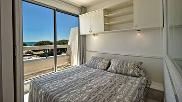 Une chambre avec une grande fenêtre, un lit recouvert d’une literie à motifs, une armoire blanche et une vue sur la plage et la mer.