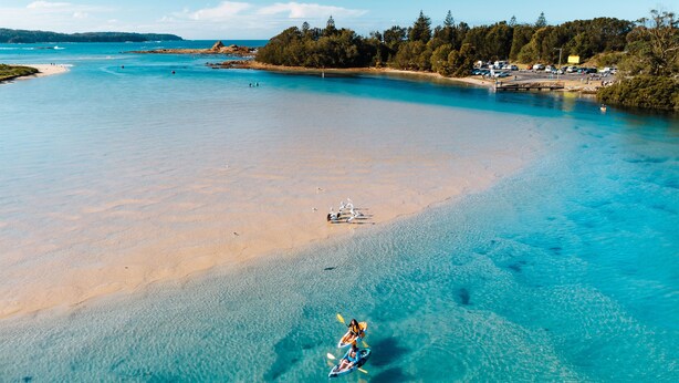 An aerial view of a beach with clear turquoise waters, a sandy shore, and a small boat with two people paddling. - The Oaks Ranch (Mossy Point)